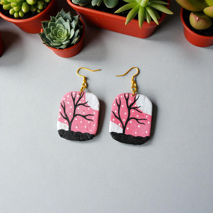 Pair of earrings with pink and white design on a gray surface with potted plants in the background.
