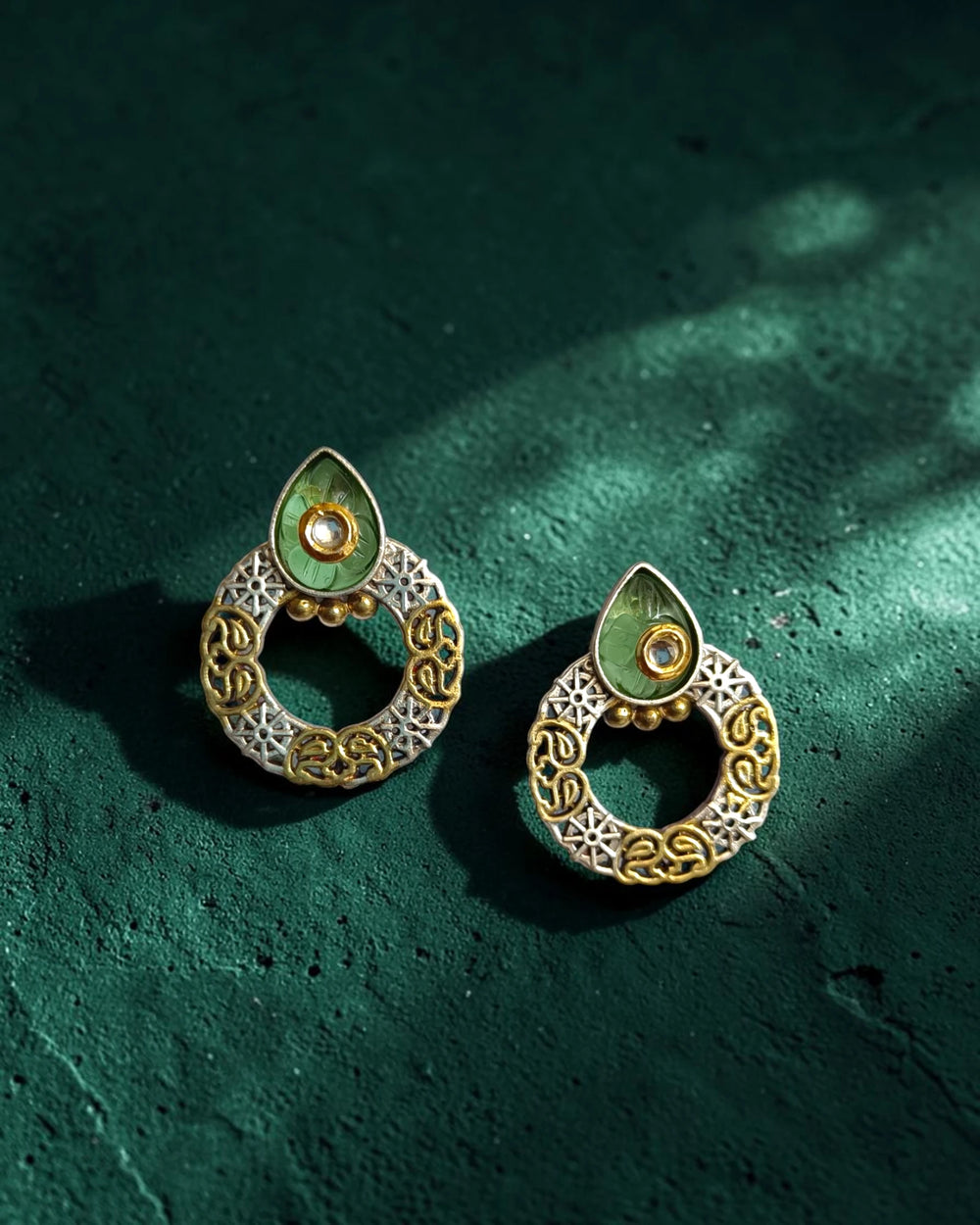 Pair of gold and silver earrings with green stones on a dark green textured surface
